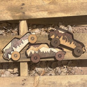 May include: Three wooden decorative trucks with mountain and tree designs. The trucks are made of layered wood in shades of brown and tan, with detailed wheels. The trucks are arranged on a wooden surface.