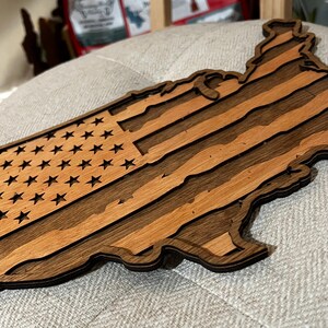 K&ouml;nnte beinhalten: Ein Holzausschnitt der Vereinigten Staaten von Amerika mit dem Design der amerikanischen Flagge. Das Holz ist dunkelbraun und hellbraun und erzeugt einen gestreiften Effekt.