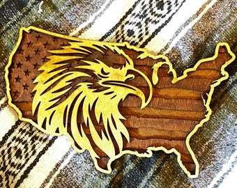 May include: Wooden wall art shaped like the United States, featuring a detailed eagle head design. The eagle is golden, set against a brown wooden background with a flag design. The piece is likely for home decor.
