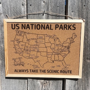 May include: A framed corkboard map of the United States with the text "US NATIONAL PARKS" and "ALWAYS TAKE THE SCENIC ROUTE". The map is etched with the names of national parks. The frame is light-colored wood, and the background is a weathered wooden surface.