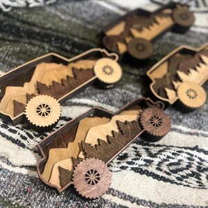 May include: Four wooden toy trucks with mountain and tree cutouts inside. The trucks have wheels and are made of various shades of brown wood. The background is a patterned blanket.
