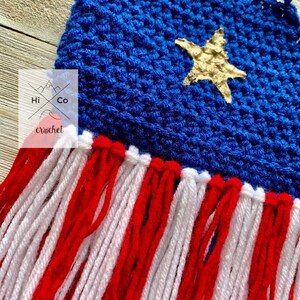 Puede incluir: Un tapiz de pared de ganchillo hecho a mano con una parte superior azul con una estrella dorada y flecos rojos y blancos. El tapiz de pared tiene el texto "Hi Co crochet". La pieza est&aacute; dise&ntilde;ada para colgarse en una pared.