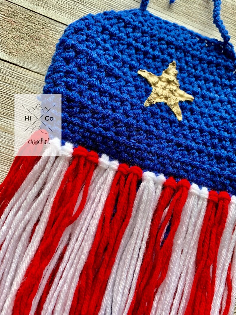 Puede incluir: Un tapiz de pared de ganchillo hecho a mano con una parte superior azul con una estrella dorada y flecos rojos y blancos. El tapiz de pared tiene el texto "Hi Co crochet". La pieza est&aacute; dise&ntilde;ada para colgarse en una pared.