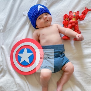 May include: A baby wearing a blue crocheted Captain America hat, blue shorts, and holding a red and yellow toy robot. A red, white and blue Captain America shield is on the white sheet.