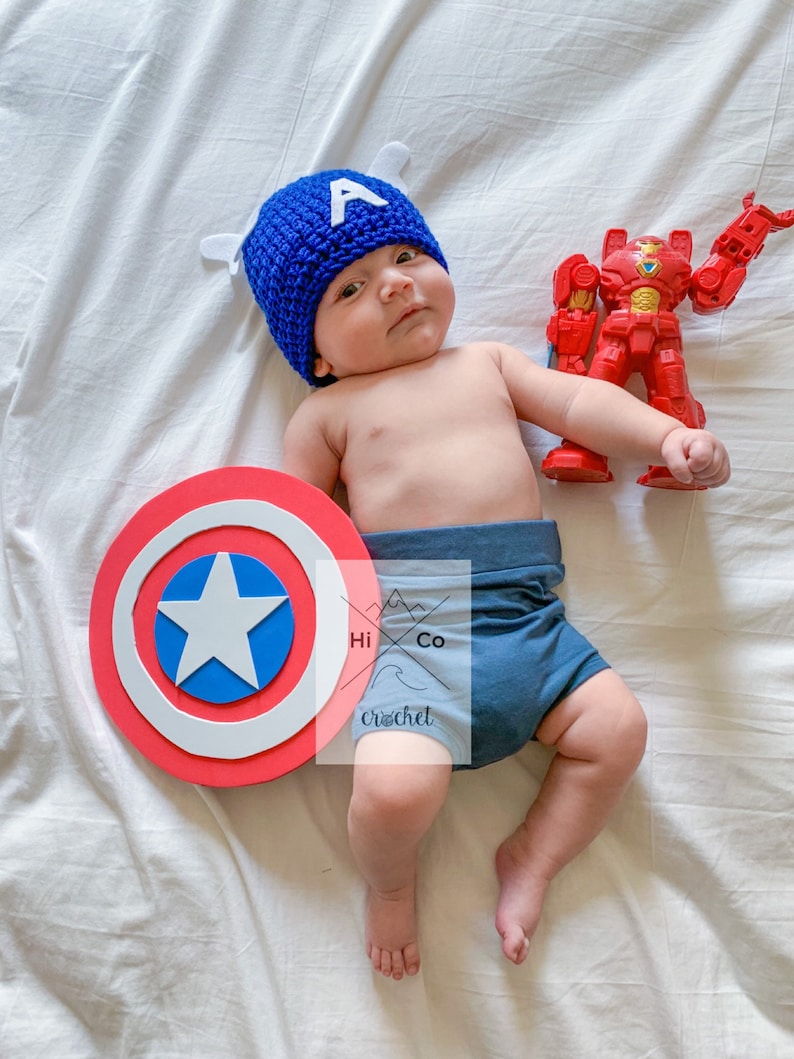 May include: A baby wearing a blue crocheted Captain America hat, blue shorts, and holding a red and yellow toy robot. A red, white and blue Captain America shield is on the white sheet.