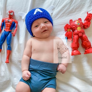 May include: A baby wearing a blue crocheted Captain America hat with white wings and a blue nappy. The baby is surrounded by superhero toys, including a red and yellow Spiderman and a red robot.