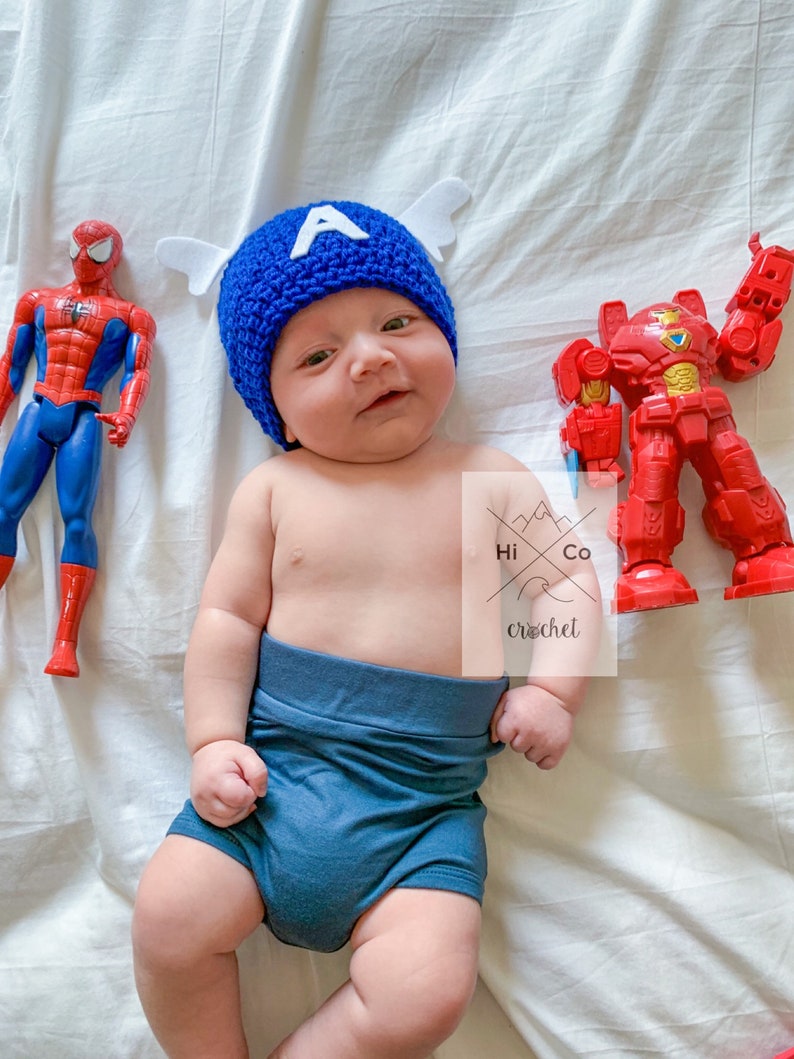 May include: A baby wearing a blue crocheted Captain America hat with white wings and a blue nappy. The baby is surrounded by superhero toys, including a red and yellow Spiderman and a red robot.