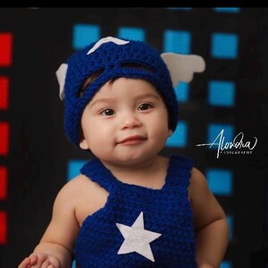 May include: A baby wearing a blue and white crocheted Captain America costume with a white star on the front. The baby is looking at the camera and has a happy expression on their face.