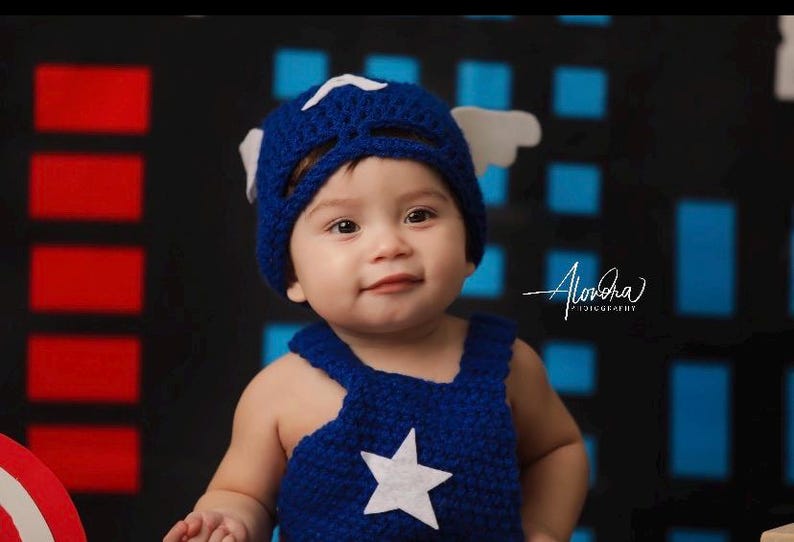 May include: A baby wearing a blue and white crocheted Captain America costume with a white star on the front. The baby is looking at the camera and has a happy expression on their face.