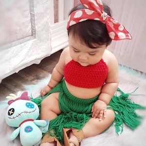 May include: A baby wearing a red crochet top and a green crochet skirt with fringe. The baby is wearing a red and white polka dot headband. The baby is sitting on a white rug with a blue and white plush toy.