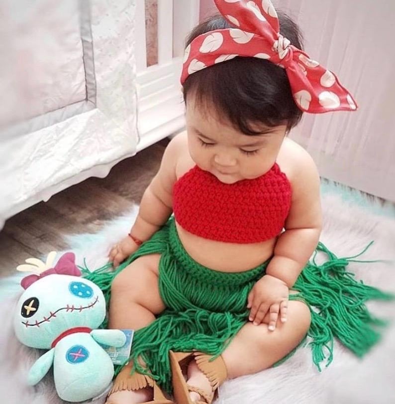 May include: A baby wearing a red crochet top and a green crochet skirt with fringe. The baby is wearing a red and white polka dot headband. The baby is sitting on a white rug with a blue and white plush toy.