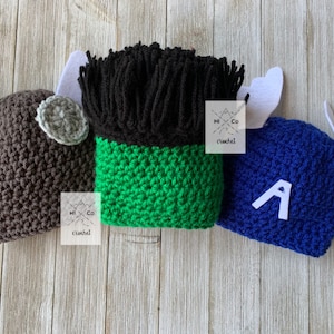 May include: Three crocheted character hats: a brown hat with gray accents, a green and black hat, and a blue hat with a white "A". Each hat has white felt accents. The hats are displayed on a light wood surface.