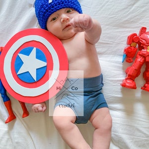 May include: A baby wearing a blue and white crocheted Captain America hat and holding a red, white and blue Captain America shield. The baby is lying on a white blanket.