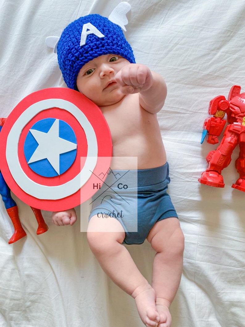 May include: A baby wearing a blue and white crocheted Captain America hat and holding a red, white and blue Captain America shield. The baby is lying on a white blanket.