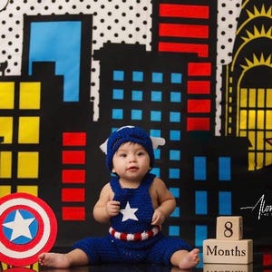 May include: A baby dressed as Captain America sits in front of a city skyline backdrop. The baby is wearing a blue and red crocheted outfit with a white star on the chest. The baby is holding a red, white, and blue Captain America shield. The backdrop is a black and white comic book style city skyline with red and yellow accents. There are wooden blocks in the foreground that say "8 Months".