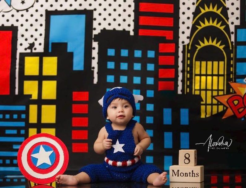 May include: A baby dressed as Captain America sits in front of a city skyline backdrop. The baby is wearing a blue and red crocheted outfit with a white star on the chest. The baby is holding a red, white, and blue Captain America shield. The backdrop is a black and white comic book style city skyline with red and yellow accents. There are wooden blocks in the foreground that say "8 Months".