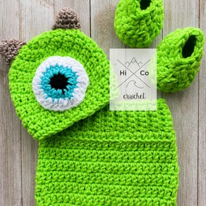 May include: Green crocheted hat and booties with a large eye design. The hat has two horns and the booties are slipper style. The set is labeled "Hi Co crochet".