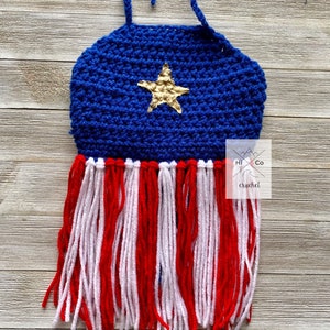 Puede incluir: Top de ganchillo hecho a mano en colores patri&oacute;ticos. El top presenta un corpi&ntilde;o azul con una estrella dorada y flecos rojos y blancos. El top est&aacute; dise&ntilde;ado con un cuello halter y se muestra sobre una superficie de madera.