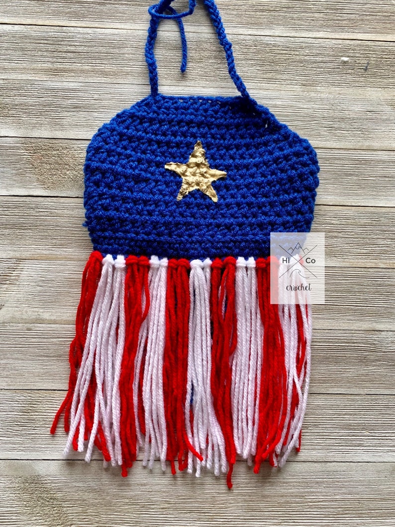 Puede incluir: Top de ganchillo hecho a mano en colores patri&oacute;ticos. El top presenta un corpi&ntilde;o azul con una estrella dorada y flecos rojos y blancos. El top est&aacute; dise&ntilde;ado con un cuello halter y se muestra sobre una superficie de madera.