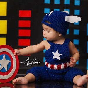 May include: A baby dressed as Captain America, wearing a blue crocheted hat and matching overalls with a red, white, and blue stripe. The baby is holding a red, white, and blue toy shield.  The background is a blue, red, and yellow city skyline.  The image also includes wooden blocks that say "8 Months".