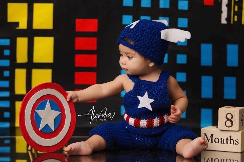 May include: A baby dressed as Captain America, wearing a blue crocheted hat and matching overalls with a red, white, and blue stripe. The baby is holding a red, white, and blue toy shield.  The background is a blue, red, and yellow city skyline.  The image also includes wooden blocks that say "8 Months".