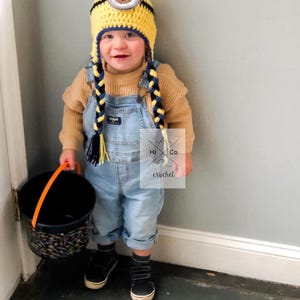 May include: A child wearing a yellow and blue crocheted Minion hat with a single large eye and braided yarn tails. The child is holding an orange-handled black bucket. Hi Co Crochet is printed on the bottom right corner of the image.