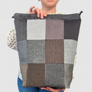 May include: A large, patchwork tote bag with a rectangular shape. The bag features a variety of grey, brown, and mauve squares. The bag is held up by a person. The bag has a drawstring closure at the top.