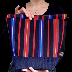 May include: A black tote bag with vertical stripes in red, yellow, blue, and white. The bottom of the bag is a solid navy blue, ribbed fabric. The bag is held up by a person.