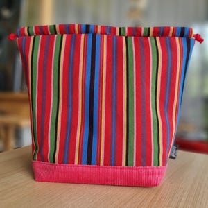 Op de afbeelding: Een stoffen trekkoordtas met een levendig gestreept patroon in rood, groen, blauw, geel en zwart. De onderkant van de tas is roze corduroy. De tas staat op een houten oppervlak.