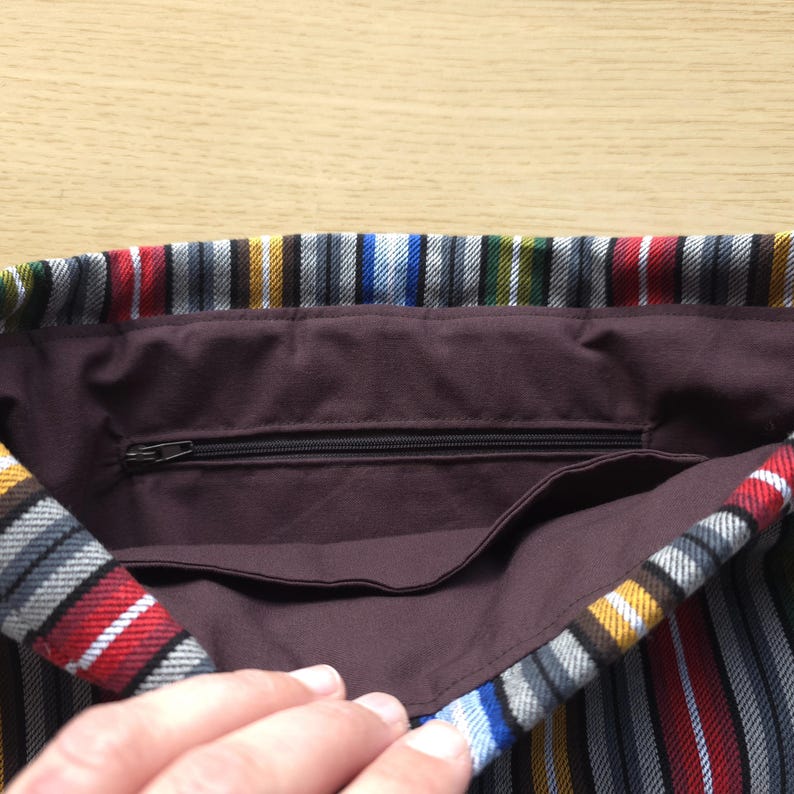 May include: A close-up view of a striped fabric bag with a dark brown interior. The bag features a black zipper and a pocket. The exterior fabric has vertical stripes in various colors, including red, yellow, blue, and gray.