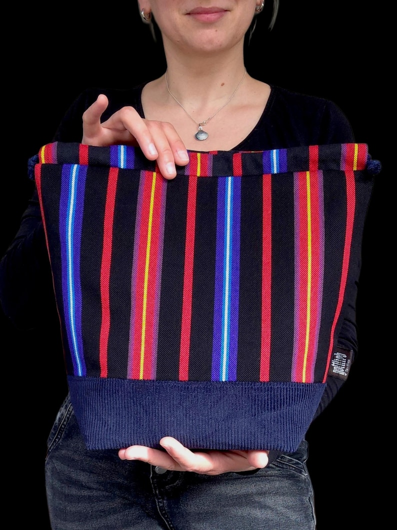 K&ouml;nnte beinhalten: Eine schwarze Tragetasche mit vertikalen Streifen in Rot, Gelb, Blau und Wei&szlig;. Der Boden der Tasche ist aus einfarbigem, marineblauem Rippstoff. Die Tasche wird von einer Person gehalten.