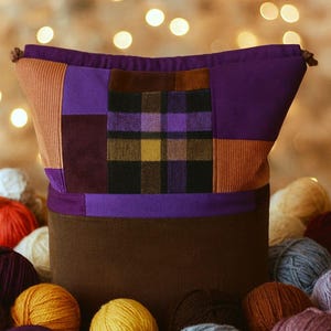 Könnte beinhalten: Eine Patchwork-Garnaufbewahrungstasche in Lila-, Braun- und Orangetönen. Die Tasche hat einen Cordboden und einen Kordelzugverschluss. Verschiedenfarbige Wollknäuel umgeben die Tasche und schaffen eine gemütliche Szene.
