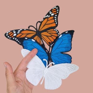 Puede incluir: Tres parches bordados de mariposas en naranja, azul y blanco. La mariposa naranja tiene detalles en blanco y negro en sus alas. La mariposa azul es de color liso. La mariposa blanca está hecha de un material similar al encaje.