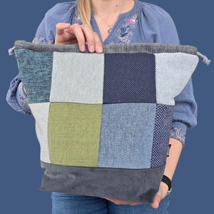 May include: A patchwork tote bag with a drawstring closure. The bag features a variety of textured fabric squares in shades of blue, green, and gray. The bottom of the bag is a solid gray fabric. The bag is held by a person.