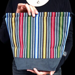 May include: A colorful tote bag with vertical stripes in red, blue, yellow, green, and gray. The bottom of the bag is a solid gray fabric. The bag is held up by a person.