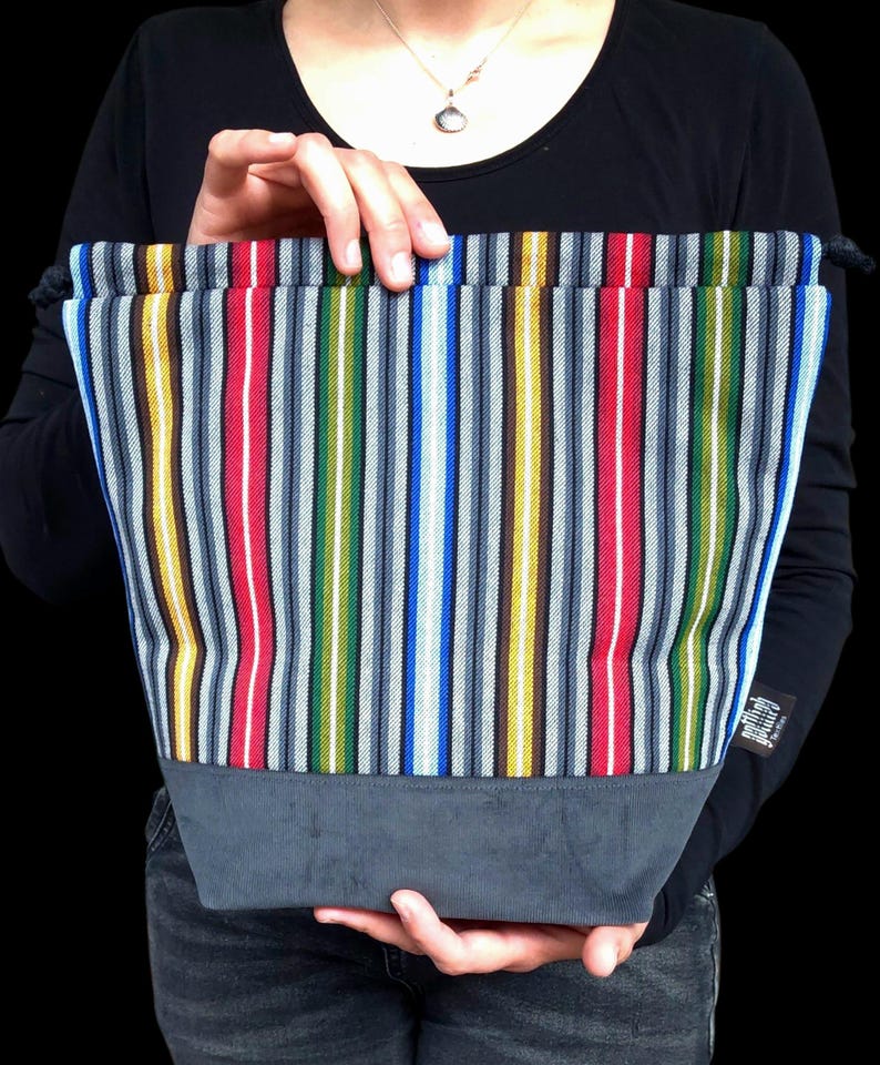 May include: A colorful tote bag with vertical stripes in red, blue, yellow, green, and gray. The bottom of the bag is a solid gray fabric. The bag is held up by a person.