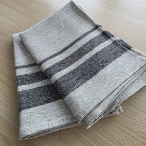 May include: Two folded linen napkins with a natural beige background and dark gray stripes. The napkins are made of a textured fabric and are neatly folded, suggesting they are ready for use.