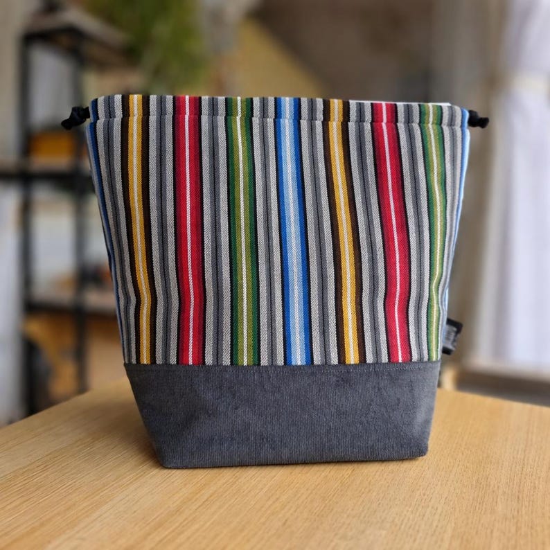 May include: A fabric drawstring bag with a gray bottom and a colorful striped pattern. The stripes include red, yellow, green, blue, and white. The bag is sitting on a wooden surface.