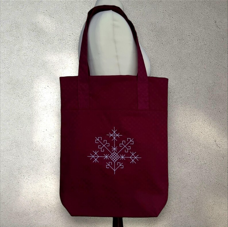 May include: A claret tote bag featuring a white snowflake design. The bag has a quilted texture and strong handles. The snowflake is embroidered on the front. The bag is designed for carrying items.
