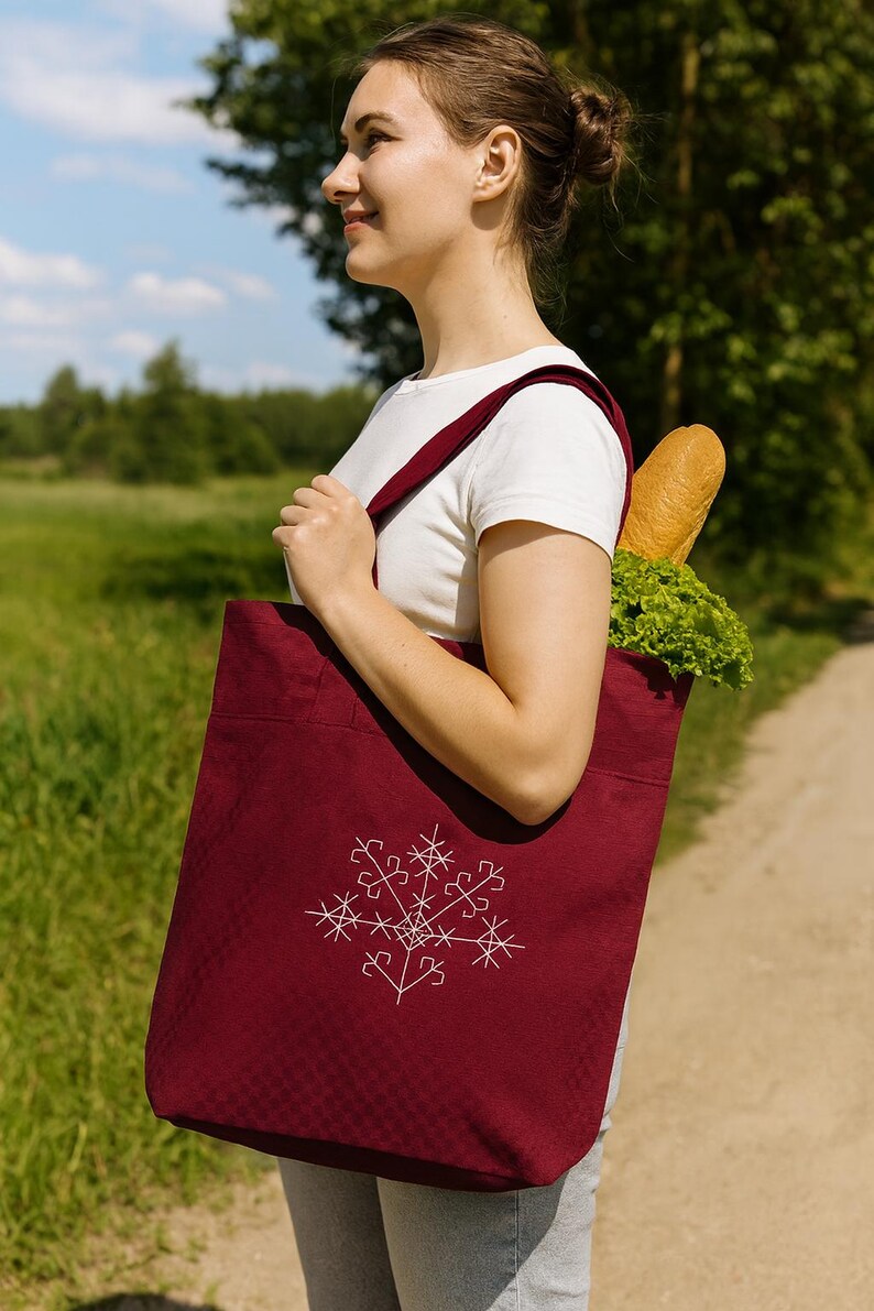 May include: A burgundy tote bag with a white embroidered snowflake design. The bag has long straps and is filled with a loaf of bread and lettuce. The person is wearing a white t-shirt.