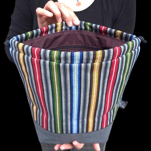 May include: A colorful striped drawstring bag with vertical stripes in red, white, gray, green, blue, yellow, and brown. The bag has a gray base and a dark brown interior. The bag is held open, revealing the inside.
