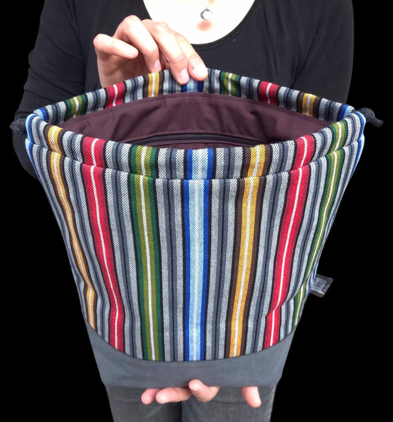 May include: A colorful striped drawstring bag with vertical stripes in red, white, gray, green, blue, yellow, and brown. The bag has a gray base and a dark brown interior. The bag is held open, revealing the inside.