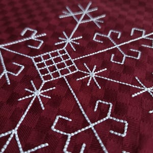 May include: Close-up of a burgundy fabric featuring a white embroidered snowflake design. The snowflake has a geometric pattern with starburst and square elements. The fabric has a subtle checkered texture.