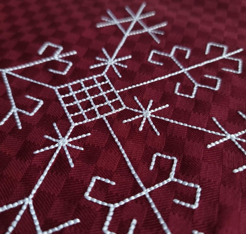 May include: Close-up of a burgundy fabric featuring a white embroidered snowflake design. The snowflake has a geometric pattern with starburst and square elements. The fabric has a subtle checkered texture.