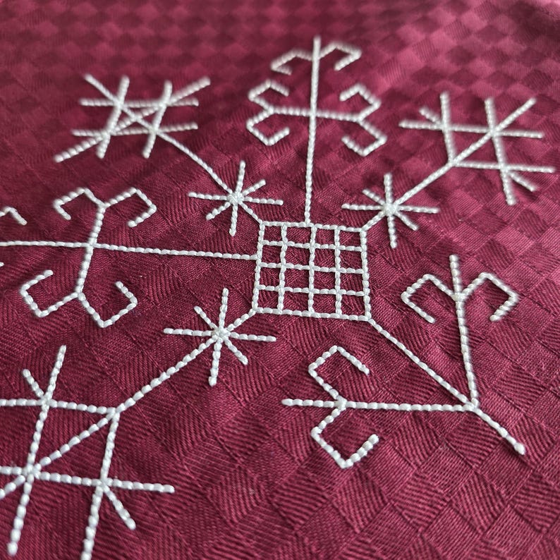 May include: Close-up of a burgundy fabric with a white embroidered snowflake design. The snowflake features geometric shapes and starbursts, creating a detailed and textured pattern. The fabric has a subtle checkered pattern.