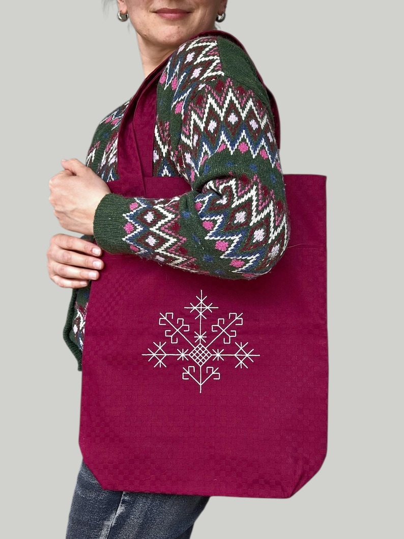 May include: A claret tote bag featuring a white geometric snowflake design. The bag has a textured surface and a rectangular shape. The tote bag is carried over the shoulder.