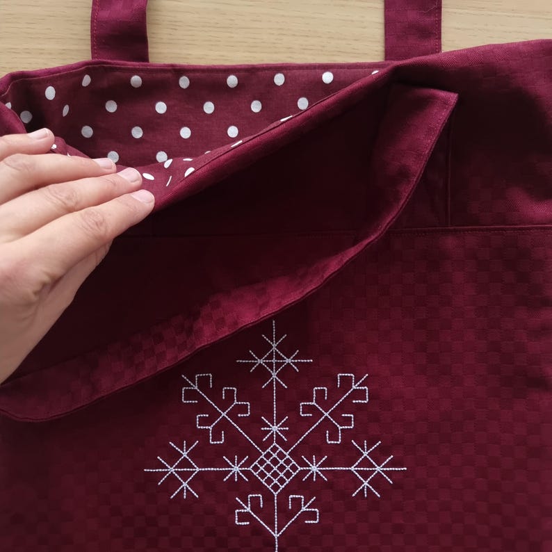 May include: Burgundy tote bag with white snowflake embroidery. The bag features a checkered pattern on the outside and a white polka dot lining. It has two handles and a large pocket. The bag is made of fabric.