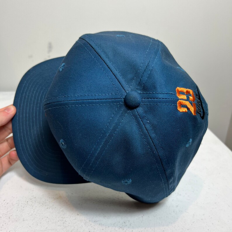 May include: A navy blue baseball cap with a curved brim and a stitched design. The cap has a small orange and white logo with the number "23" on the side.