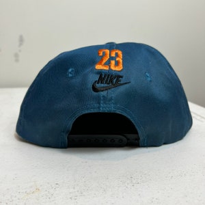 May include: A blue baseball cap with a black Nike swoosh logo and the number 23 embroidered in orange on the back.