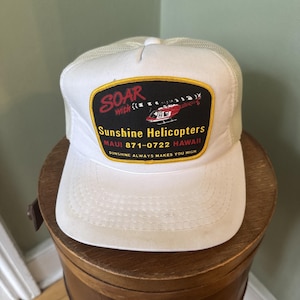 May include: White and beige trucker cap with a curved peak. The front has a patch reading "SOAR with Sunshine Helicopters" and a red helicopter graphic. The text "MAUI 871-0722 HAWAII" is also visible.